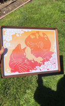 Risograph print in bring orange and red ink of a person petting a cat, they both lean their heads into one another. Piece is framed and held in someone's hand against grass.