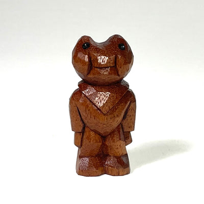 Small whittled wood carving of a frog with chubby cheeks, standing with its arms to its side. It wears clothing and a bandana around its neck.