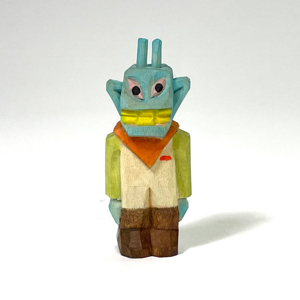 Painted whittled wood sculpture of a blue alien with a goofy yellow smile and crooked eyes. It stands with its arms to its side, wearing work clothes with an orange bandana around its neck.