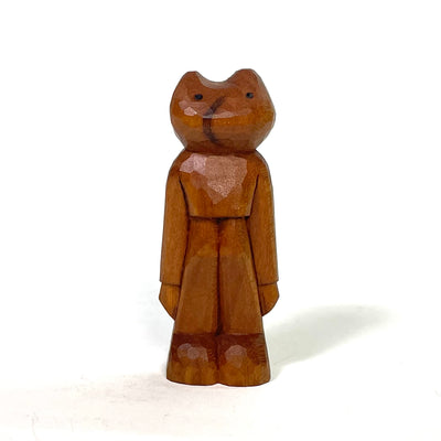Small unpainted whittled wood sculpture of a frog like person, standing tall with its long arms to its side. It wears a sweater, pants and boots.