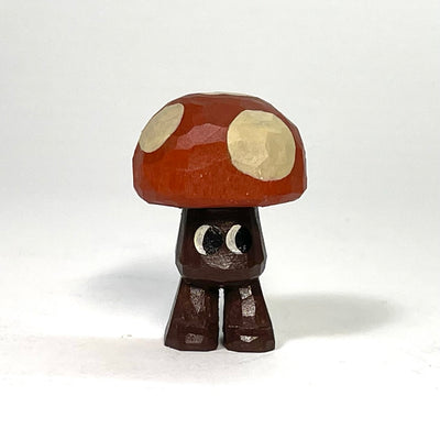 Painted whittled wood sculpture of a small mushroom, with a rounded red cap with white dots. It has a small, thick brown body with only a pair of legs and a set of eyes looking off to the side.