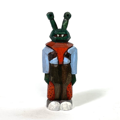 Painted whittled wood sculpture of a dark green alien, with eyes like a snail. It stands on 2 legs with its arms to its side and wears a blue jacket with a red shirt and brown pants, with red patches.