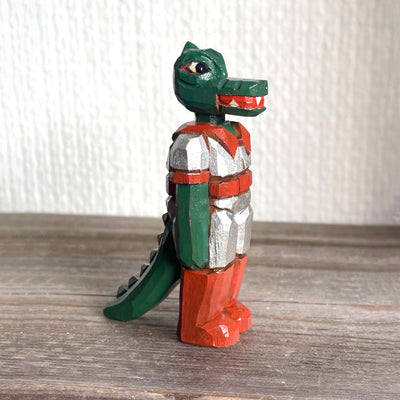 Whittled wooden sculpture of a green dinosaur, akin to a crocodile, wearing a space suit of silver and red.