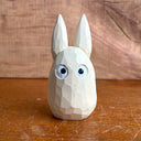Whittled wooden sculpture of a small, off white ghost creature with large cartoon eyes, tall pointed ears and no limbs or other facial features. It looks straight ahead.