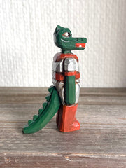 Whittled wooden sculpture of a green dinosaur, akin to a crocodile, wearing a space suit of silver and red.