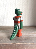 Whittled wooden sculpture of a green dinosaur, akin to a crocodile, wearing a space suit of silver and red.