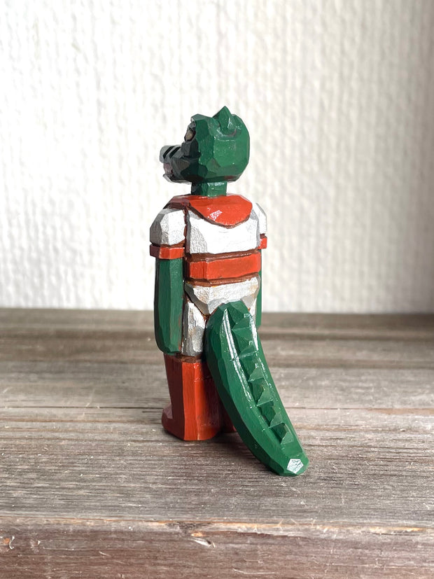 Whittled wooden sculpture of a green dinosaur, akin to a crocodile, wearing a space suit of silver and red.
