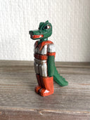 Whittled wooden sculpture of a green dinosaur, akin to a crocodile, wearing a space suit of silver and red.