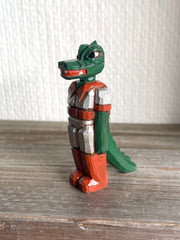 Whittled wooden sculpture of a green dinosaur, akin to a crocodile, wearing a space suit of silver and red.