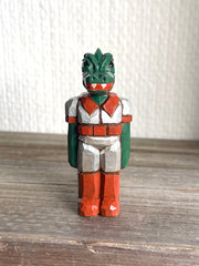 Whittled wooden sculpture of a green dinosaur, akin to a crocodile, wearing a space suit of silver and red.