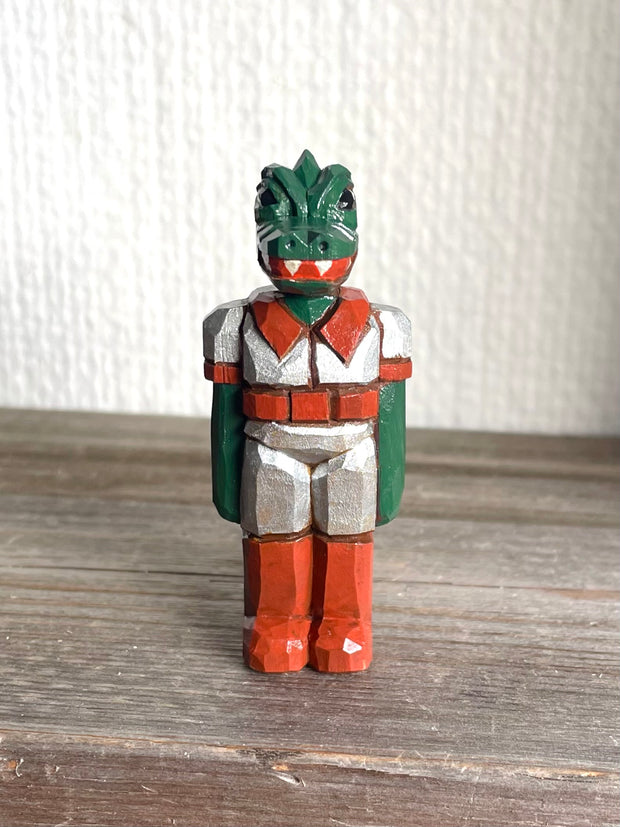 Whittled wooden sculpture of a green dinosaur, akin to a crocodile, wearing a space suit of silver and red.