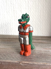 Whittled wooden sculpture of a green dinosaur, akin to a crocodile, wearing a space suit of silver and red.