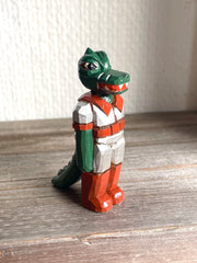 Whittled wooden sculpture of a green dinosaur, akin to a crocodile, wearing a space suit of silver and red.