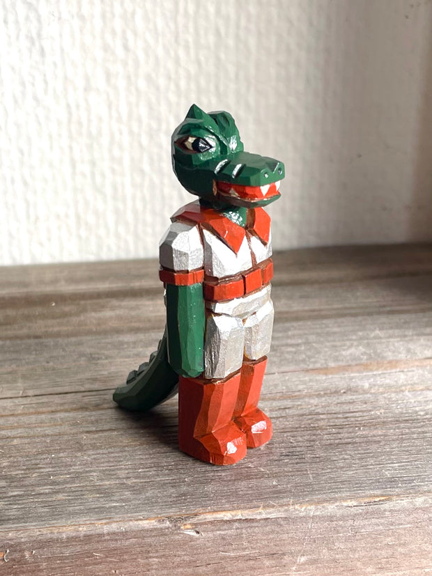 Whittled wooden sculpture of a green dinosaur, akin to a crocodile, wearing a space suit of silver and red.