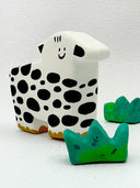 Clay sculpture of a stylistic cow, with a pointed head and a very simple smiley face. 3 sculpted clumps of grass surround it.