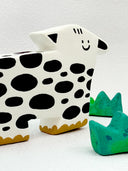 Clay sculpture of a stylistic cow, with a pointed head and a very simple smiley face. 3 sculpted clumps of grass surround it.