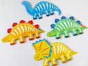 Four colorful dinosaur-shaped flat artworks arranged on a plain white background. The set includes a blue long-necked dinosaur, a yellow-orange Stegosaurus with red spikes, a green Tiktaos, and a yellow Triceratops with blue spikes and bone details.