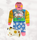 Paper cut out of a person wearing a shirt with patterning and skateboarding dog on it. Sitting at her feet is a white dog with brown and black spots.