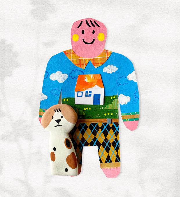 Paper cut out of a person wearing a shirt with patterning and a house on it. Sitting at her feet is a tall brown, black and white dog.
