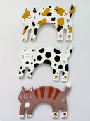 Three clay cat-shaped sculptures, laying flat, with different color patterns on a white background