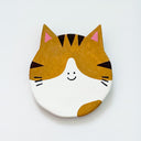 Round clay sculpture of a cat face, totally flat, with brown and white coloring and black stripes. It has a tiny smile in the middle of the face.