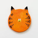 Round clay sculpture of a cat face, totally flat, with orange coloring and black stripes. It has a tiny smile in the middle of the face.