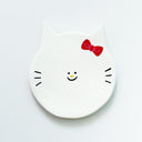 Round clay sculpture of a cat face, totally flat, with white coloring and red hair bow. It has a tiny smile in the middle of the face.