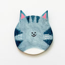 Round clay sculpture of a cat face, totally flat, with gray and white coloring and bluish gray stripes. It has a tiny smile in the middle of the face.