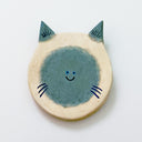 Round clay sculpture of a cat face, totally flat, with gray and white coloring and gray color spots. It has a tiny smile in the middle of the face.