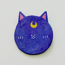 Round clay sculpture of a cat face, totally flat, with bluish purple coloring and a tiny moon on its forehead. It has a tiny smile in the middle of the face.
