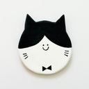Round clay sculpture of a cat face, totally flat, with black and white coloring. It has a tiny smile in the middle of the face and wears a small bowtie.