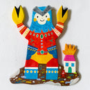 Flat colorful sculpture of a kaiju monster with crab claws for hands, blue head and red and yellow outfit. It stands on dirt near a house on fire with a tiny red devil character.