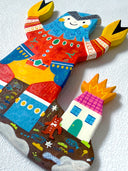 Flat colorful sculpture of a kaiju monster with crab claws for hands, blue head and red and yellow outfit. It stands on dirt near a house on fire with a tiny red devil character.