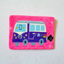 A rectangular pink decorative plaque featuring a hand-painted purple and blue smiling VW bus, displayed on a textured white wall.