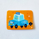 A flat artwork featuring a hand-painted illustration of a smiling blue taxi on an orange background, photographed on a textured white wall.