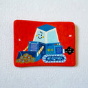 A hand-painted clay flat artwork featuring a whimsical blue bulldozer with a smiling face on a bright red background, mounted on a textured white wall.