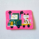 A pink flat artwork featuring a whimsical hand-painted illustration of a green truck carrying a white and brown calico cat, set against a textured white background.