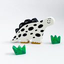 A clay sculpture of a dinosaur colored like a cow, with a white body, black spots, black spikes, and gold feet, accompanied by two small green grass pieces, photographed on a clean white background.