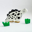A hand-painted clay dinosaur painted to look like a cow with black spots on a white body, standing next to two small green decorative plants on a white surface against a white wall.