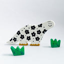 A handcrafted clay dinosaur sculpture in white with black floral patterns and yellow feet, accompanied by two small green grass pieces, photographed on a clean white background.