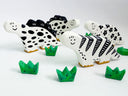 A set of hand-painted clay dinosaur sculptures, of stylized dinosaurs colored like cows and four small green grass pieces, arranged on a white surface against a white background.