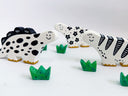 A set of hand-painted clay dinosaur sculptures, of stylized dinosaurs colored like cows and four small green grass pieces, arranged on a white surface against a white background.