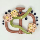 Ceramic sculpture of a brown 2 handled flat vessel shape with 3 chrysanthemum flowers attached and a green snake slithering along the main body of the vessel. Small black dots hang or stand atop the handles.