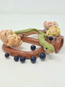 Ceramic sculpture of a brown 2 handled flat vessel shape with 3 chrysanthemum flowers attached and a green snake slithering along the main body of the vessel. Small black dots hang or stand atop the handles.