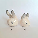 Two white ceramic figures of heads with horns - one is longer than the other.