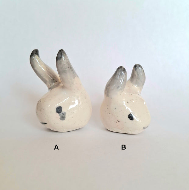 Two white ceramic figures of heads with horns - one is longer than the other.