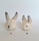 Two white ceramic figures of heads with horns - one is longer than the other.