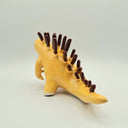 Ceramic sculpture of a orange-ish tan kaiju dinosaur creature with many thin spikes on its back, akin to a porcupine. Spikes look like they are chocolate dipped.
