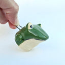 Ceramic sculpture of a green frog's head with a cream colored underside. it has a closed mouth and large, wide eyes. Attached to the top is a wire semicircle mounting element.