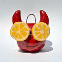 Ceramic sculpture of a red devil head, with a smooth cute face and softly curved horns. On its eyes are slices of lemons.